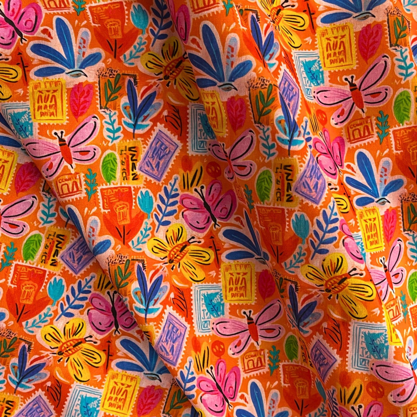 Orange Postcard Garden cotton fabric folded to show crisp woven construction and layered scattered print of postcards, botanicals and butterflies — breathable 100% cotton ideal for handmade blouses, skirts and summer dresses