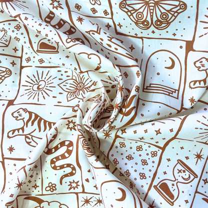 Close-up of white cotton poplin fabric showing irregular patchwork print in warm caramel and ginger tones with celestial tiger floral and star motifs — Oeko-Tex Standard 100 certified, ideal for slow fashion statement dressmaking