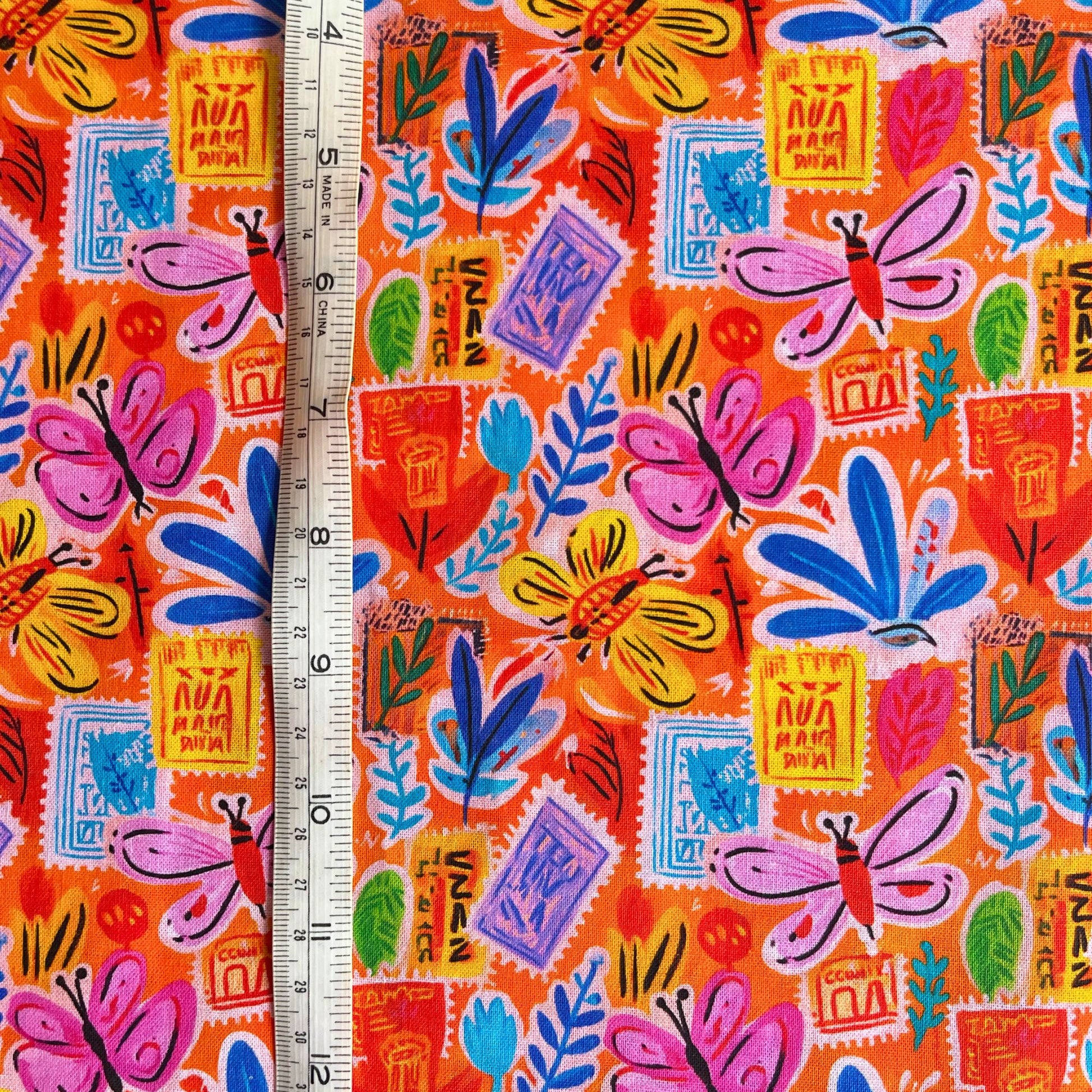 Close-up of orange cotton fabric showing scattered vintage postcard motifs, delicate botanical details and butterfly accents — crisp breathable 100% cotton woven ideal for slow fashion dressmaking and children's sewing