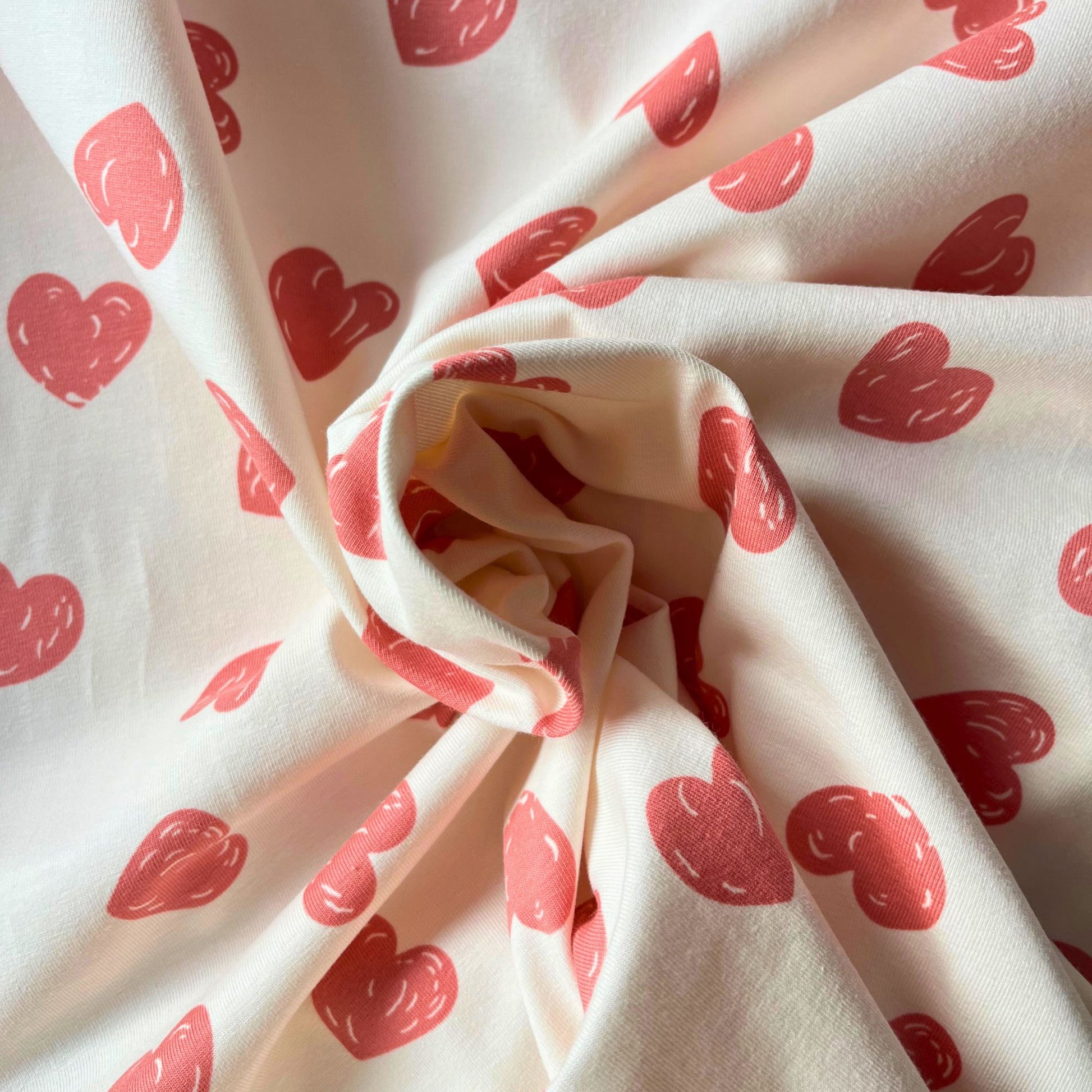 Close-up of cream stretch jersey fabric showing playful hand-drawn style rose pink doodle heart print — soft breathable cotton elastane knit ideal for handmade children's tops, leggings and babygrows