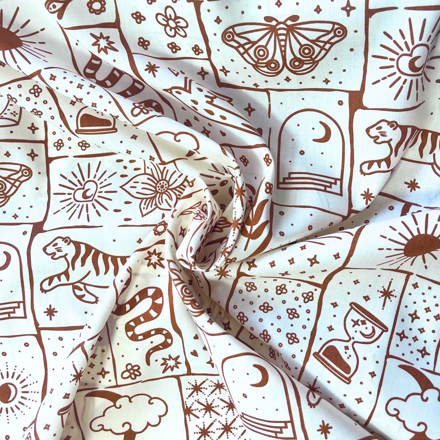 Flat lay of white base celestial patchwork poplin fabric draped to show crisp lightweight 110gsm drape — irregular caramel and ginger patches with tigers, florals and moons, perfect for handmade shirt dresses and statement co-ords