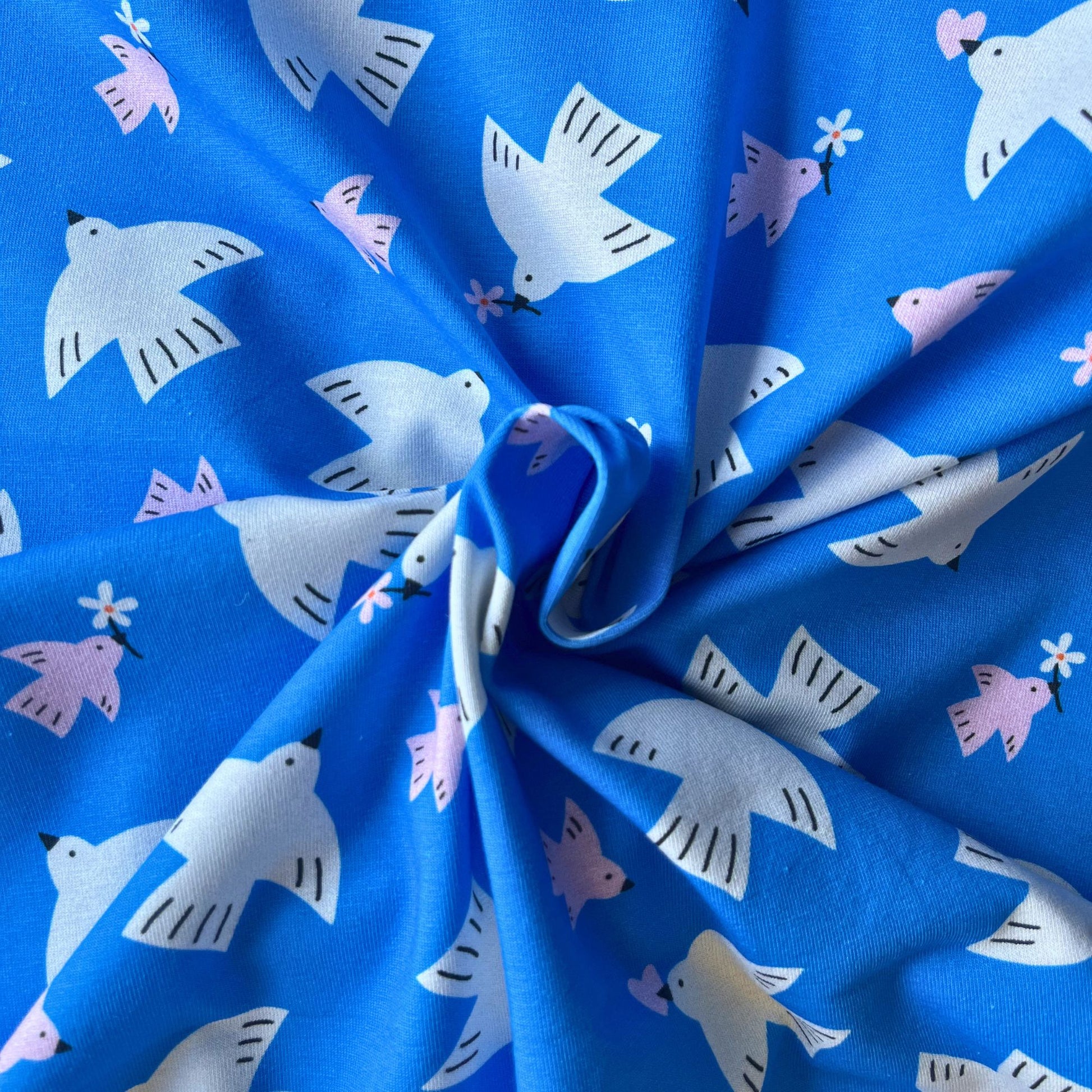 Close-up of bright blue digital cotton jersey fabric showing white and pink bird and floral print — crisp digitally printed 100% cotton knit ideal for slow fashion dressmaking and children's sewing