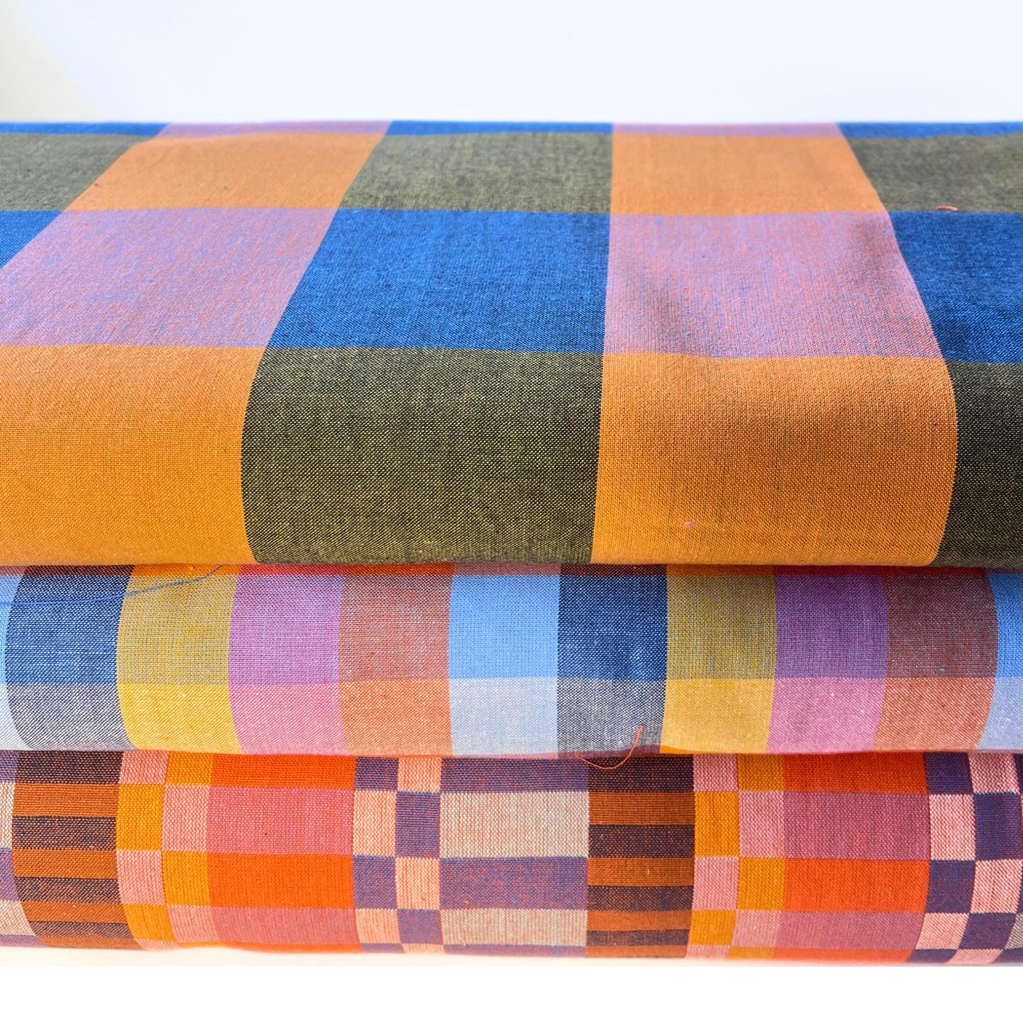 Three folded plaid fabrics stacked on top of each other, showcasing different multicolor patterns including orange, blue, green, pink, and yellow.
