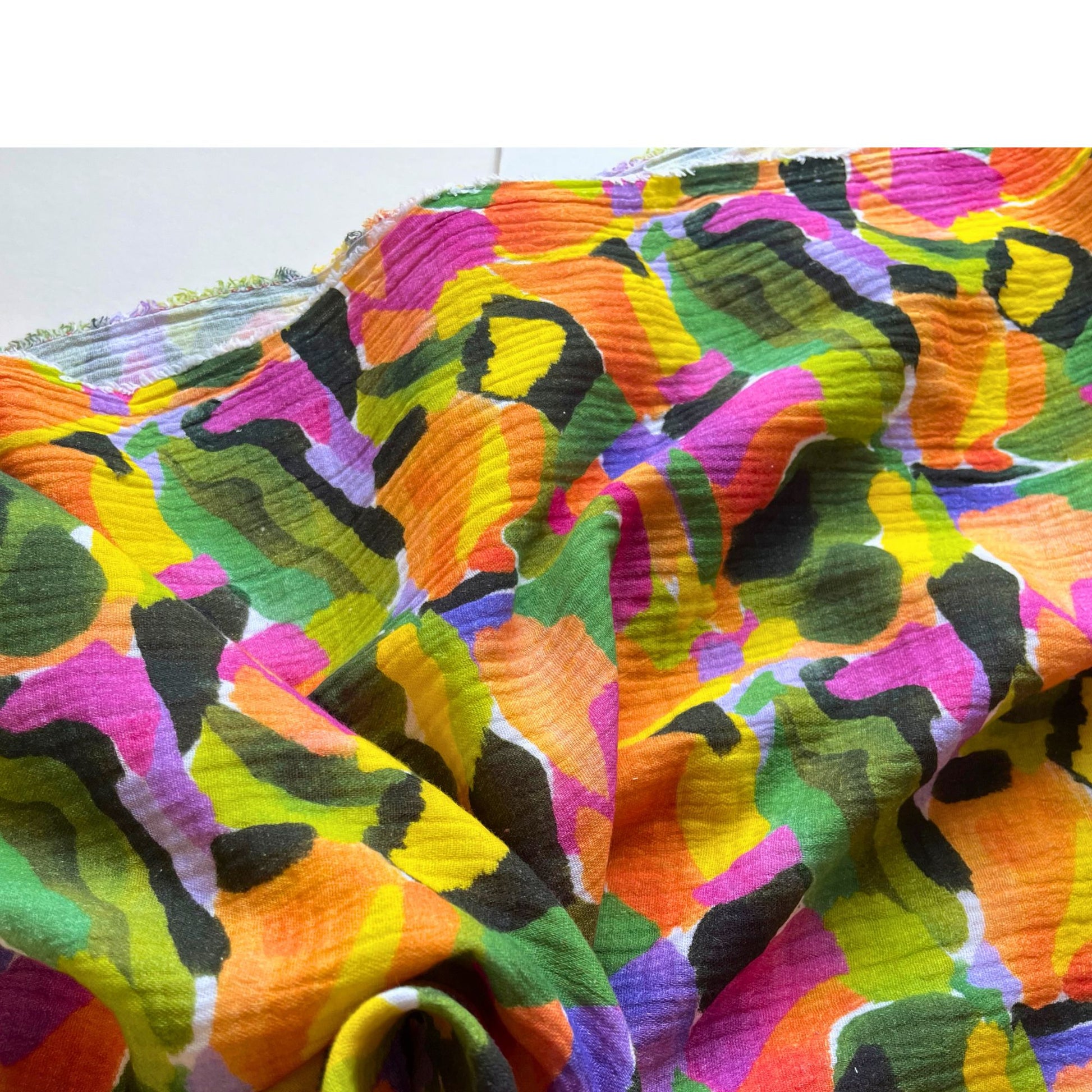 Close-up of white double gauze fabric showing vibrant painterly tropical print in green, purple and orange tones — soft two-layer 100% cotton woven ideal for handmade baby muslins, children's clothing and adult dressmaking
