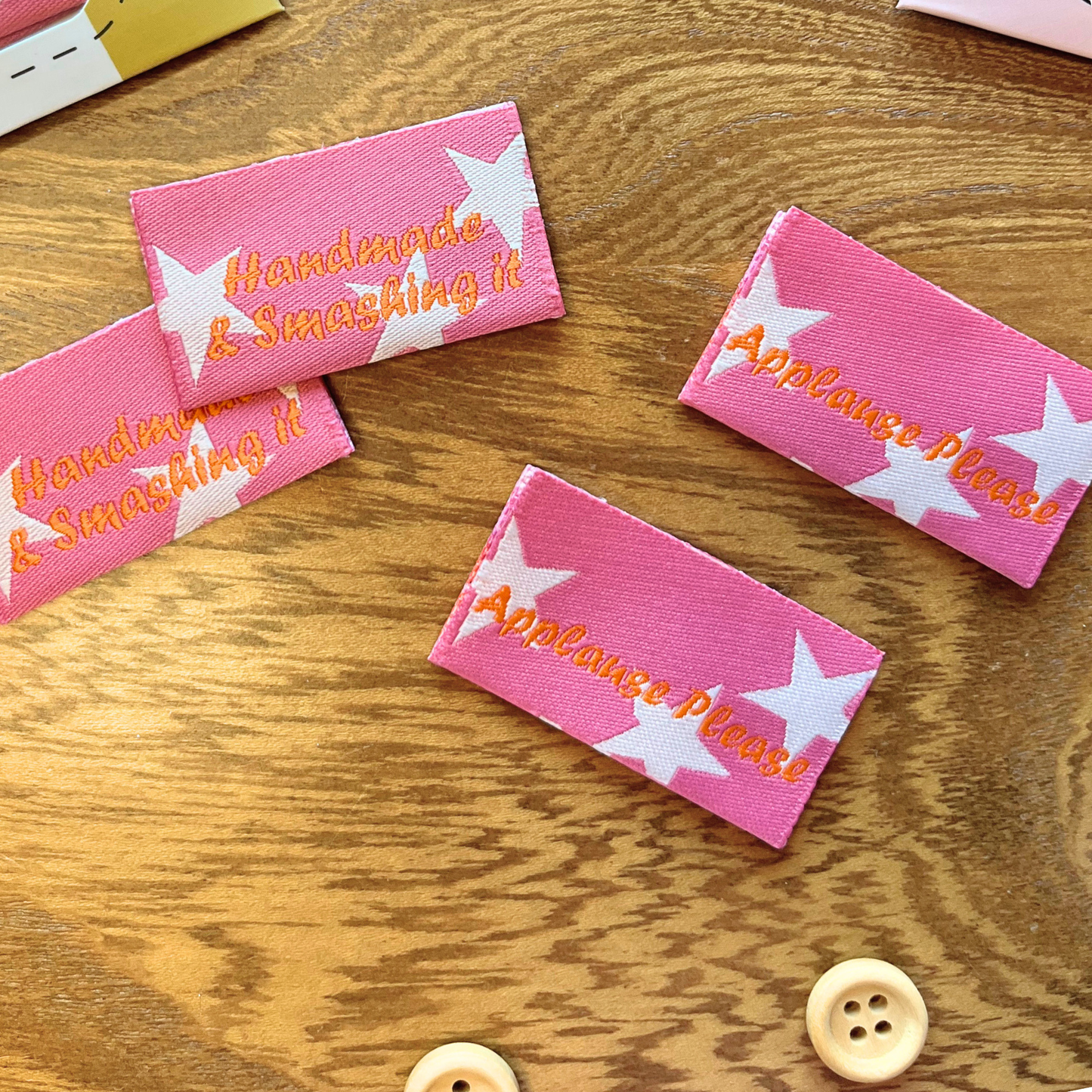 Flat lay of Handmade and Smashing It sewing labels showing pink front with stars and orange lettering alongside Applause Please reverse — recycled polyester folded labels, UK made
