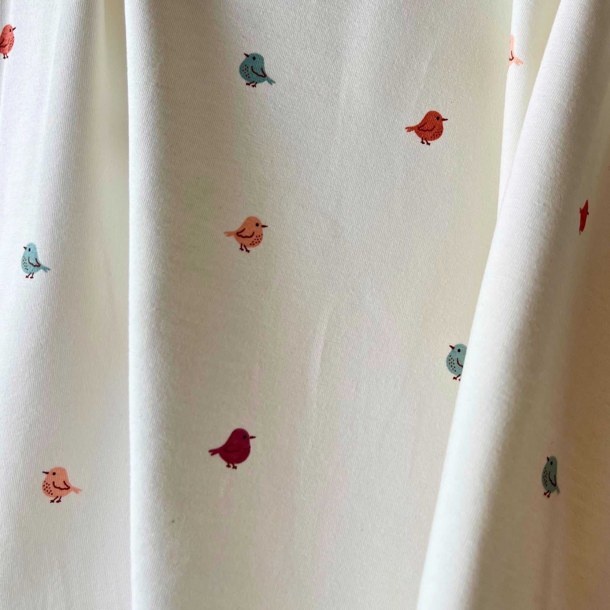 Close-up of white stretch jersey fabric showing tiny scattered bird print in muted rose, warm putty and fresh teal tones — soft breathable cotton elastane knit ideal for handmade babygrows and children's garments