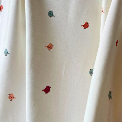 Close-up of white stretch jersey fabric showing tiny scattered bird print in muted rose, warm putty and fresh teal tones — soft breathable cotton elastane knit ideal for handmade babygrows and children's garments
