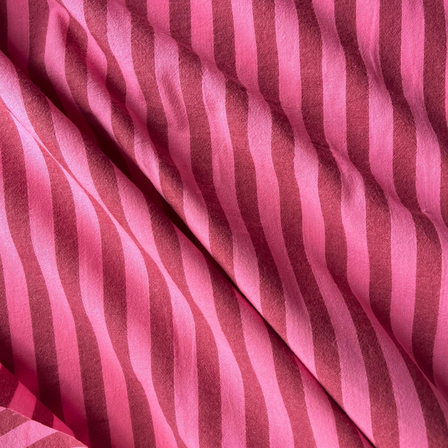 Flat lay of washed pink and red yarn dyed stripe fabric draped to show lightweight 115gsm drape and relaxed laundered finish — classic cotton stripe woven perfect for handmade summer dresses and blouses