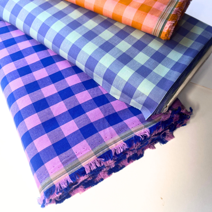 Three folded plaid fabrics stacked on a white surface, showing blue, purple, pink, and orange checkered patterns with fringed edges.