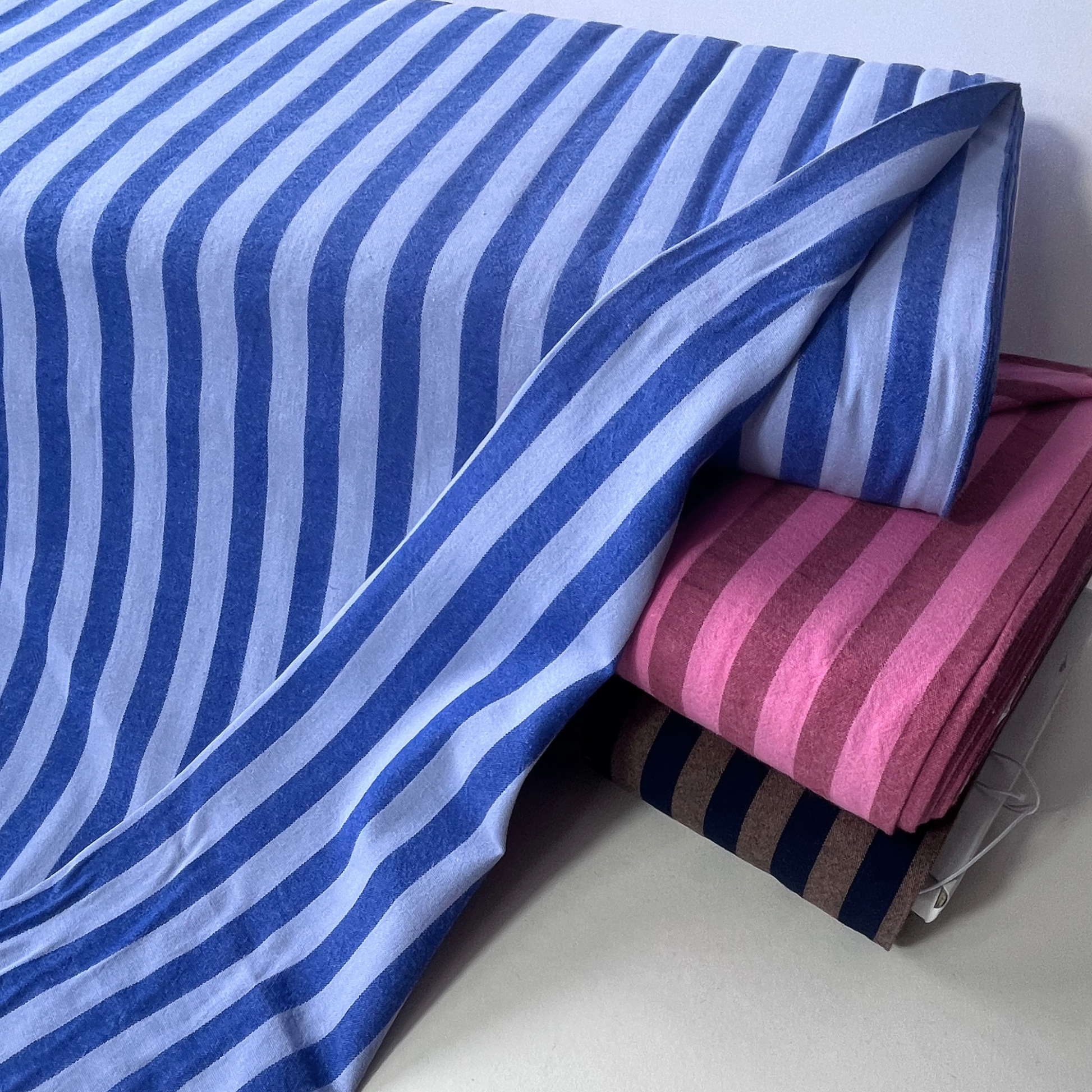 Blue and white striped fabric draped over a stack of folded striped textiles in pink, brown, and navy on a light neutral background.