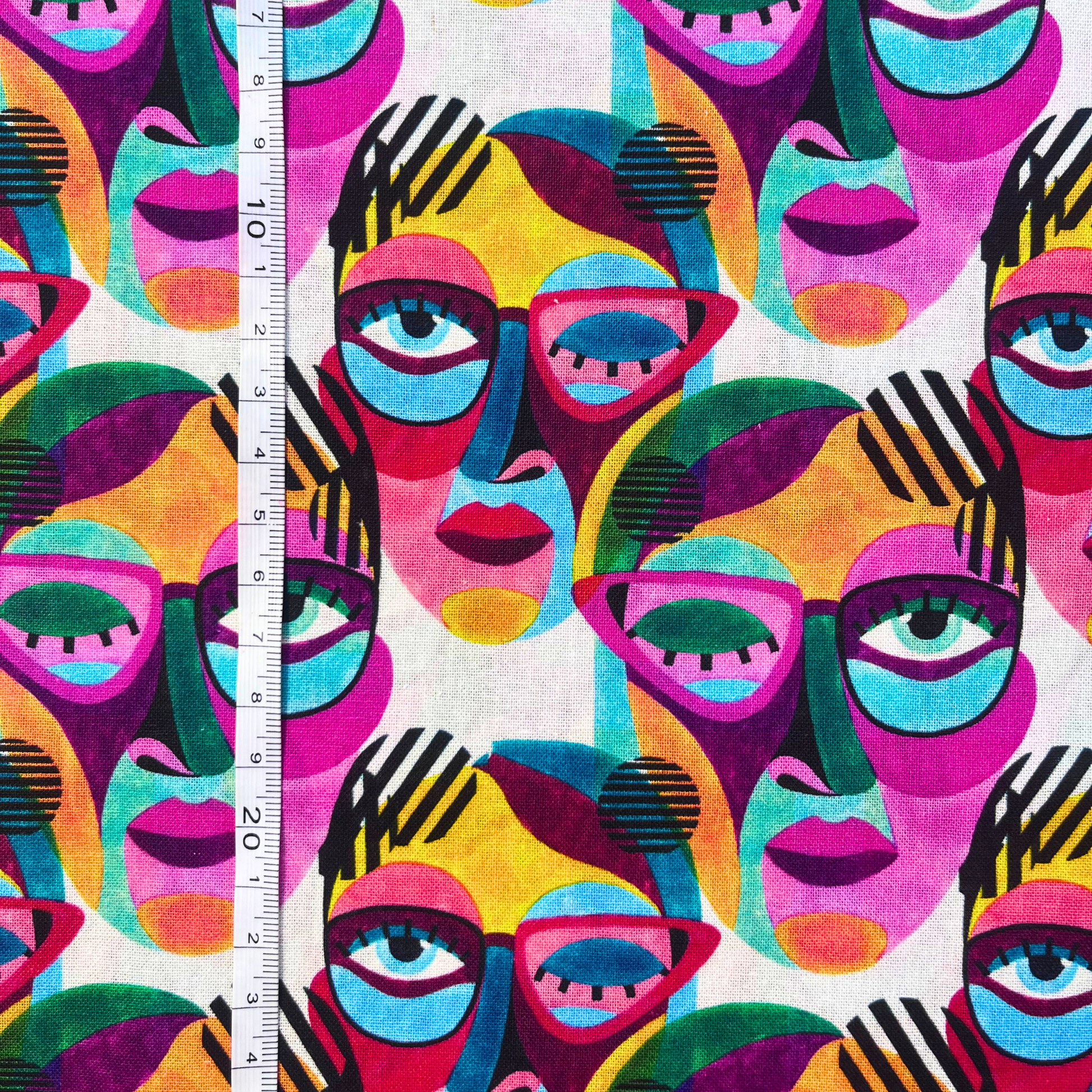 Flat lay of Little Johnny abstract painted faces cotton fabric draped to show crisp 115gsm weight and generous 149cm width — bold multicolour rainbow face print on white base perfect for handmade statement wrap dresses and oversized shirts