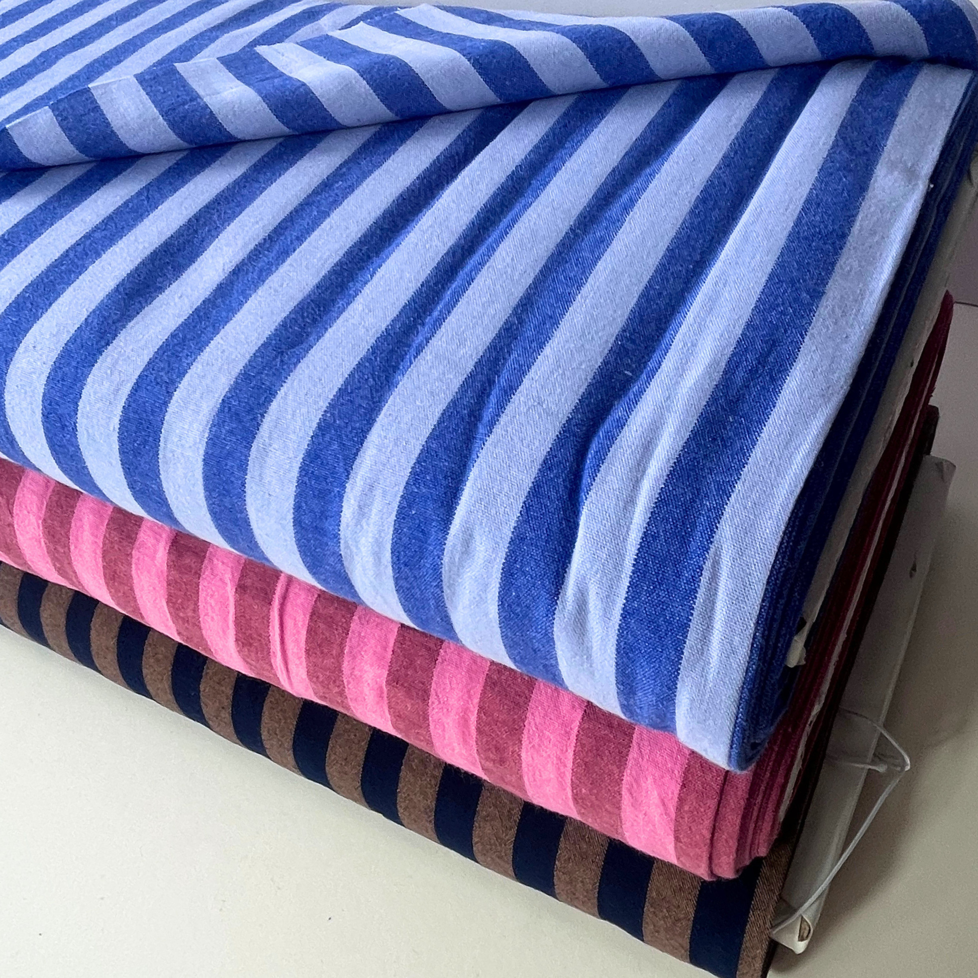 A stack of folded striped yarn dyed cotton dressmaking fabrics, with the top piece in blue stripes and the bottom pieces showing pink and brown stripes, photographed on a light neutral surface.