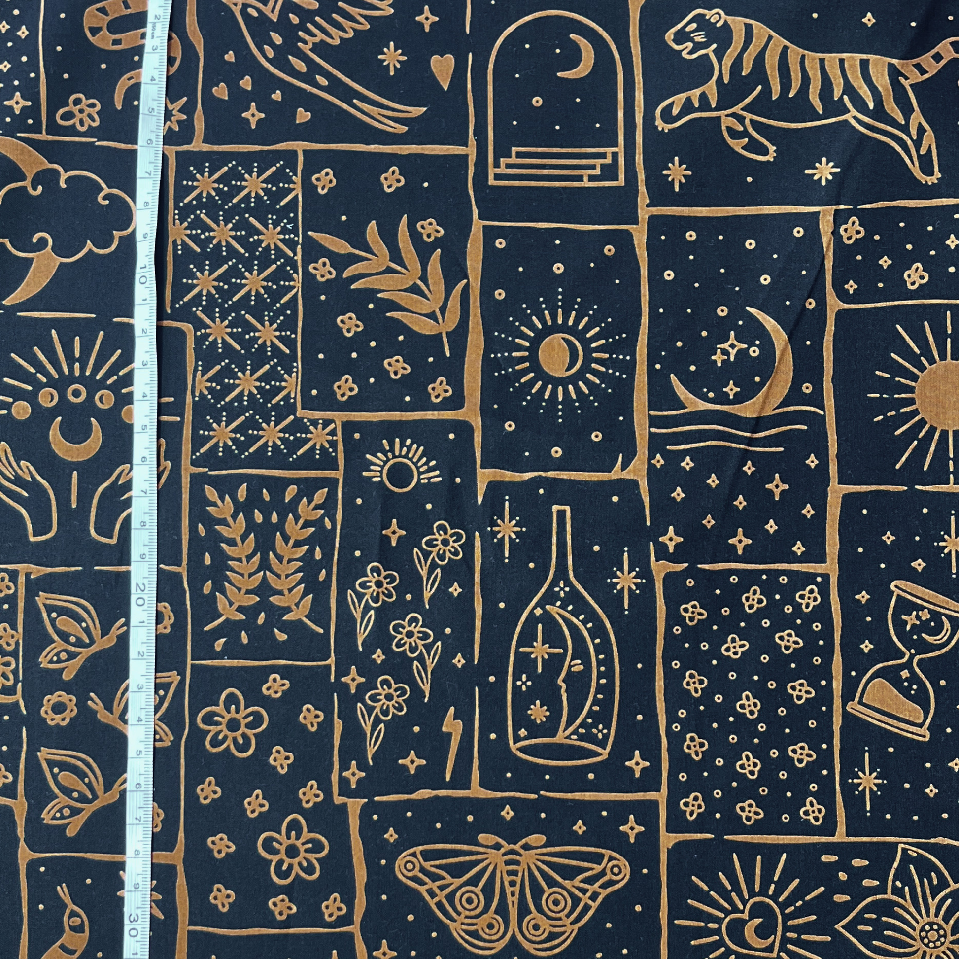 Close-up of black cotton poplin fabric showing irregular patchwork print in warm caramel and ginger tones with celestial tiger floral and star motifs — Oeko-Tex Standard 100 certified, ideal for slow fashion statement dressmaking