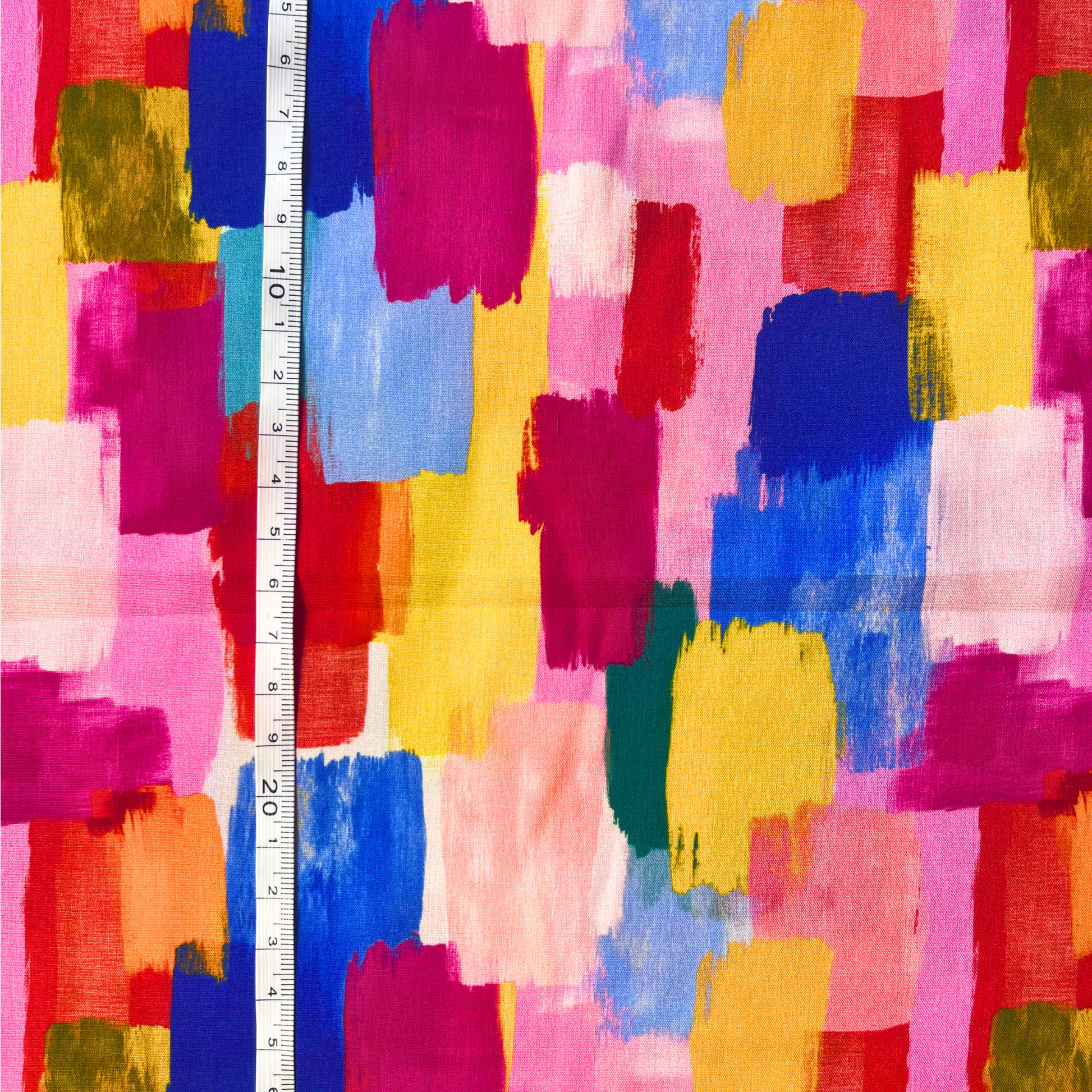 Multicolor abstract painterly pattern fabric with brushstroke blocks of pink, blue, yellow, red, orange, green, and white, shown with a white tape measure for scale.
