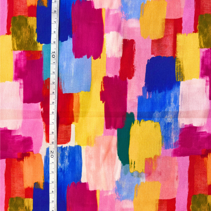 Multicolor abstract painterly pattern fabric with brushstroke blocks of pink, blue, yellow, red, orange, green, and white, shown with a white tape measure for scale.