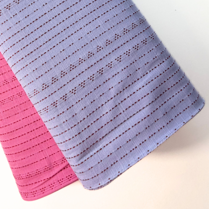 Two textured fabric cloths, one lavender with a dark red dotted pattern and one pink, layered on a light beige background.