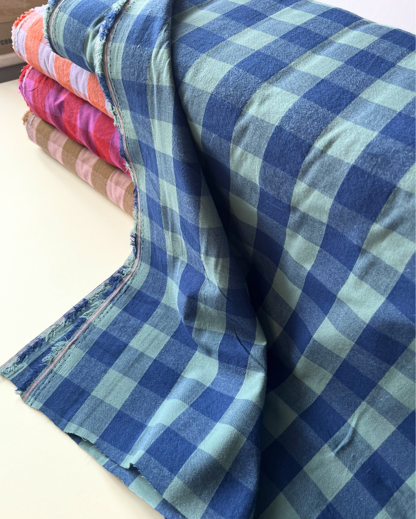 Flat lay of blue and teal gingham cotton fabric draped to show 135gsm weight and natural drape — 3cm yarn dyed check fabric perfect for handmade women's shirts, handmade beyond nine inspired dresses and summer trousers