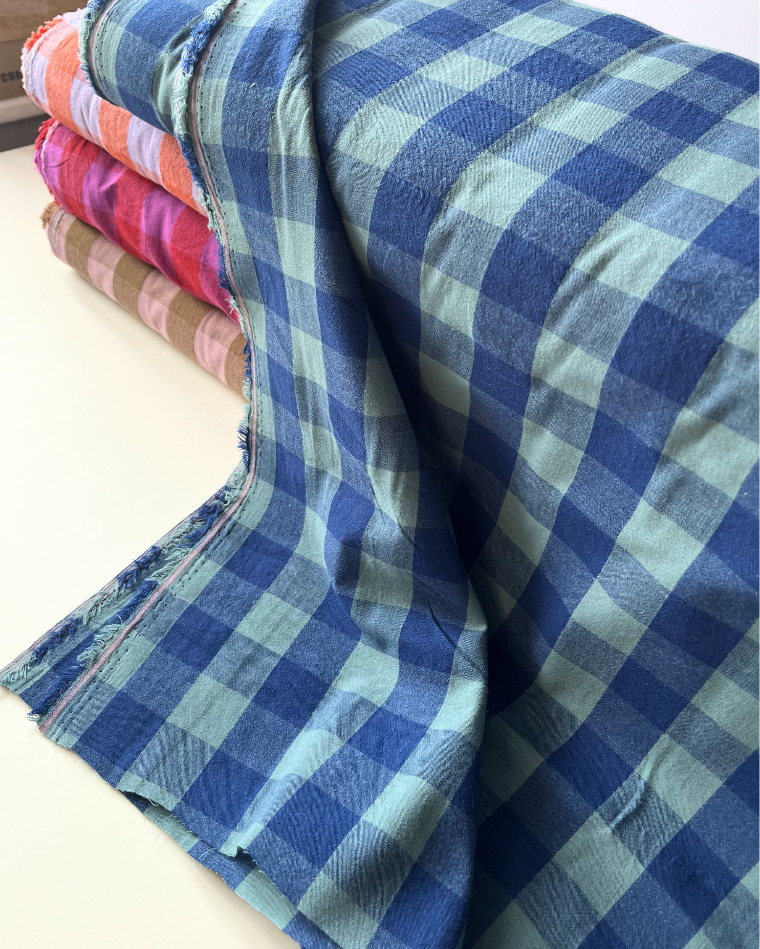 Flat lay of blue and teal gingham cotton fabric draped to show 135gsm weight and natural drape — 3cm yarn dyed check fabric perfect for handmade women's shirts, handmade beyond nine inspired dresses and summer trousers