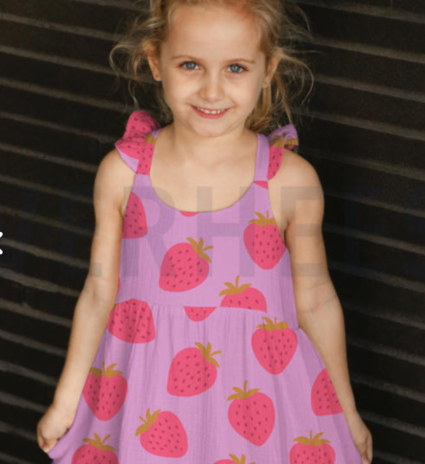 A young girl smiling and wearing a light purple sleeveless dress with a pink and red strawberry pattern, set against a dark slatted background.