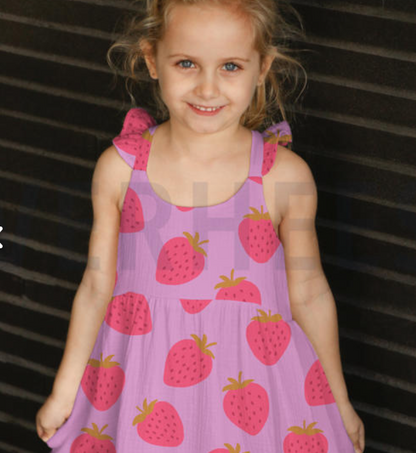 A young girl smiling and wearing a light purple sleeveless dress with a pink and red strawberry pattern, set against a dark slatted background.
