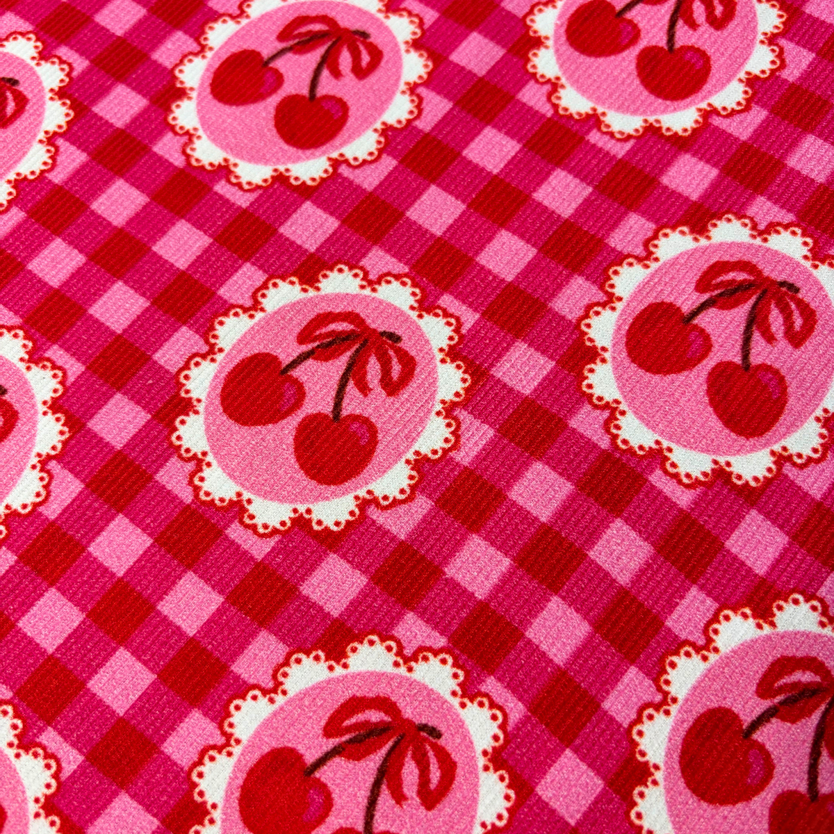Close-up of red and pink gingham cotton needlecord showing cute cherry motifs in circular frames on a repeat pattern, soft corduroy texture slow fashion dressmaking fabric UK