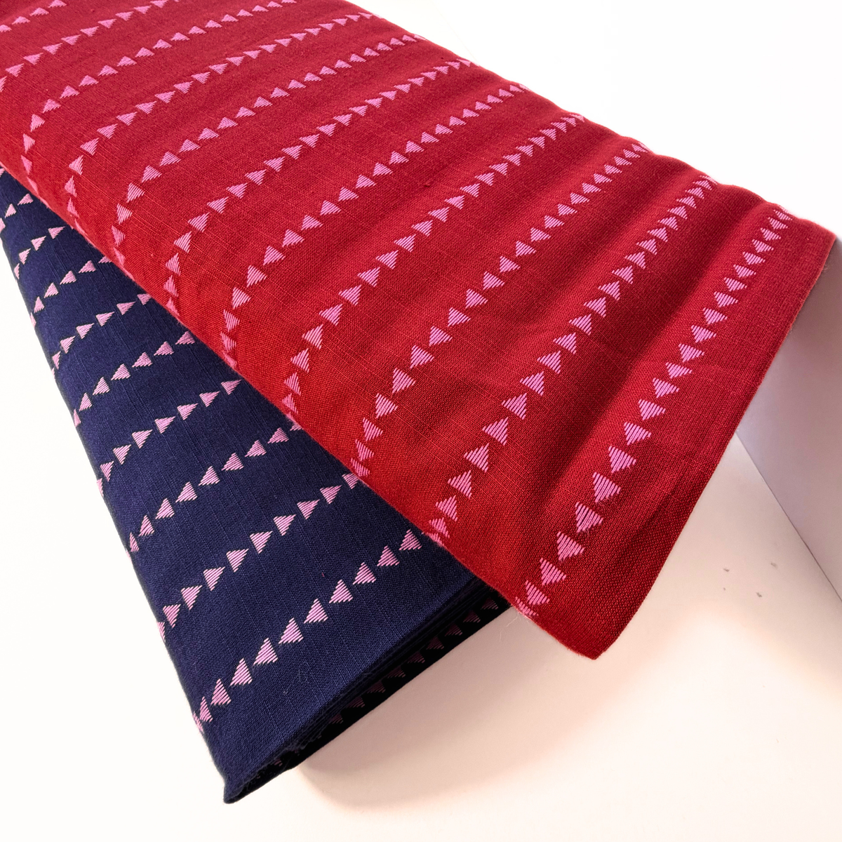 Two folded woven textiles with a repeating pink triangular dash pattern, shown on a white background. One is red and the other is navy blue.