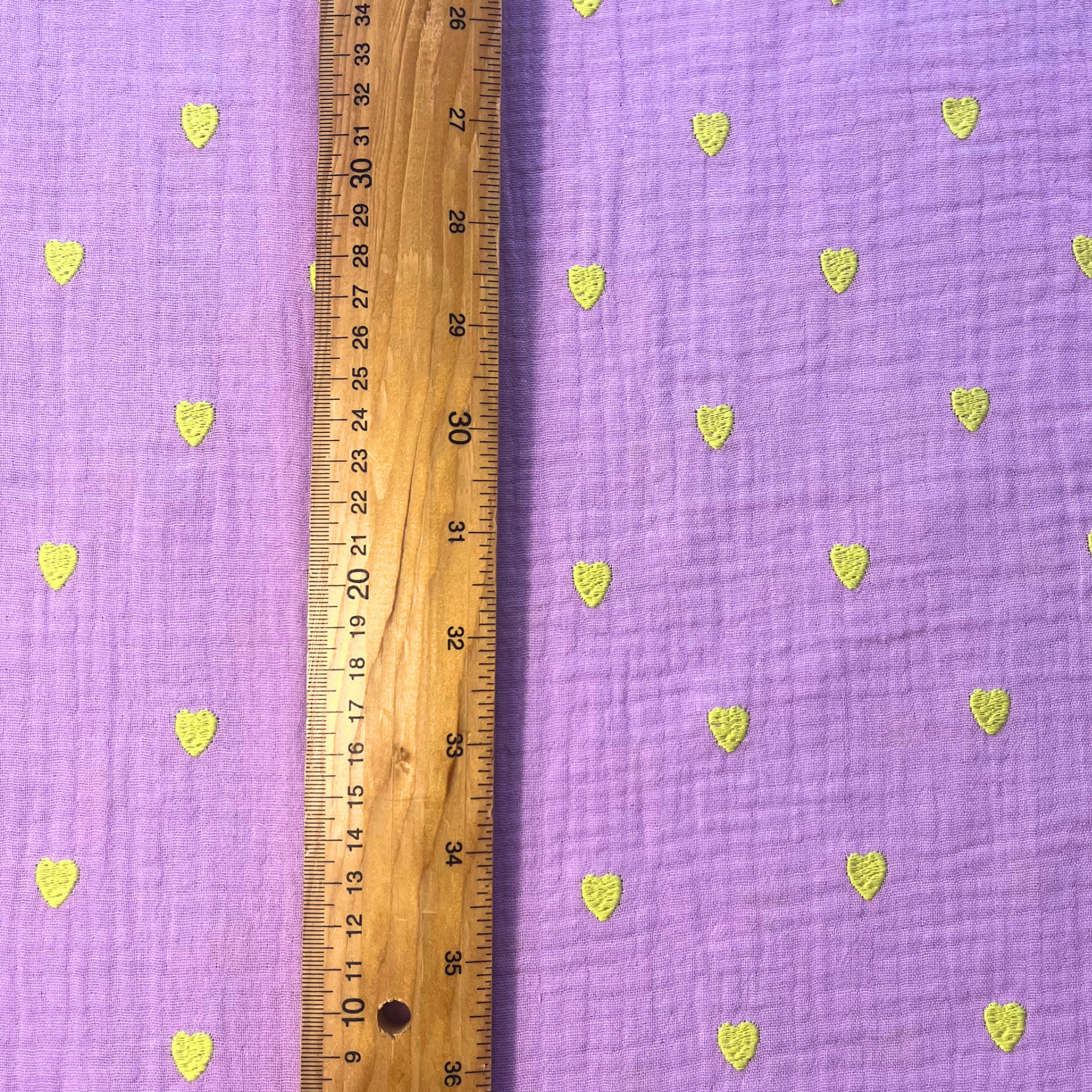 Close-up of soft lavender double gauze fabric showing tiny scattered neon yellow embroidered hearts with tactile stitched texture — Oeko-Tex certified 140gsm 100% cotton woven ideal for handmade children's dresses, baby swaddles and adult blouses