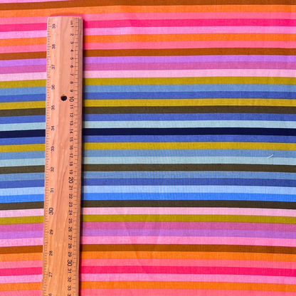 A close-up of a striped dressmaking fabric with horizontal bands of pink, orange, blue, yellow, green, and brown, shown with a wooden ruler on the left for scale.