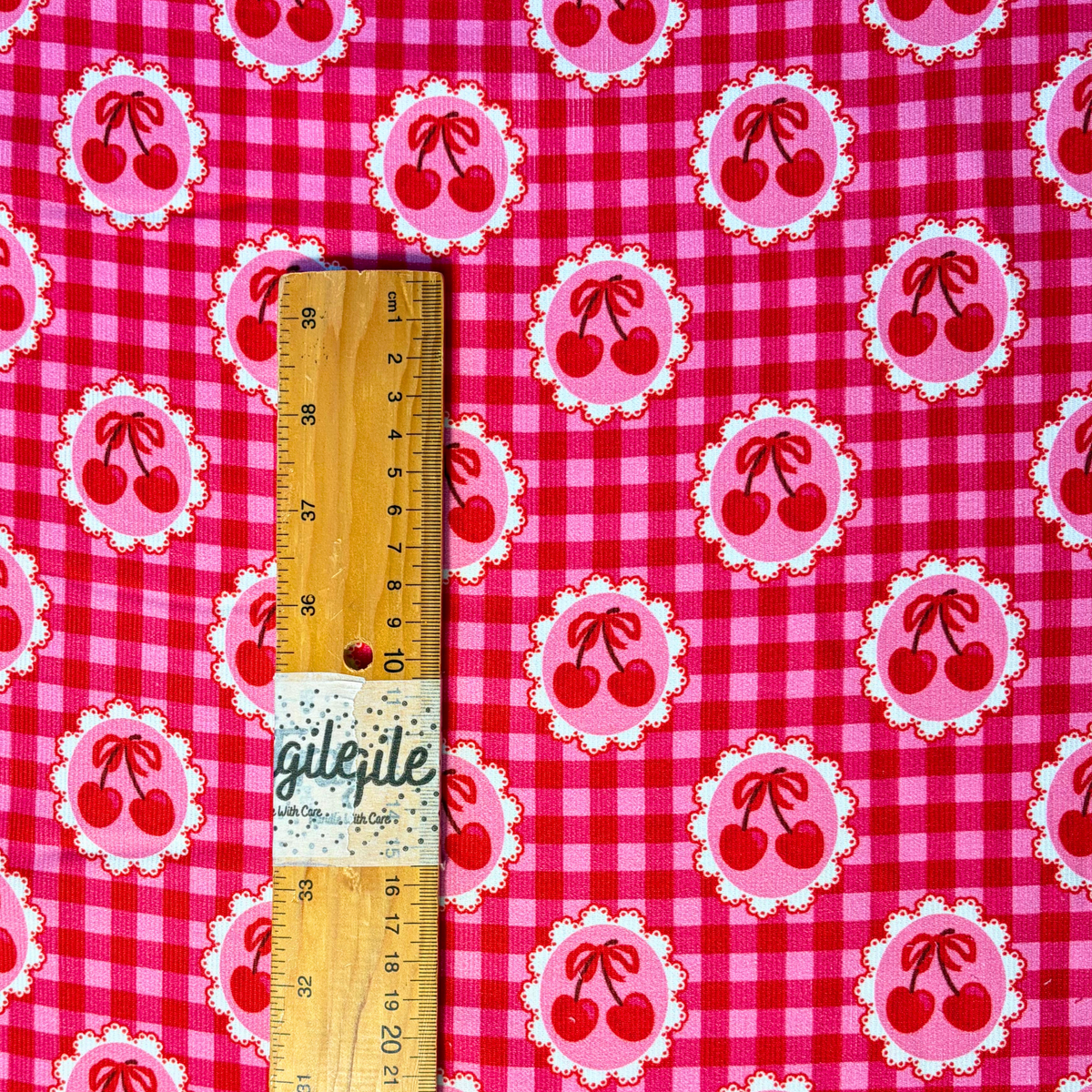Red pink cherry gingham needlecord fabric flat lay showing 125gsm drape and 112cm width, cotton corduroy with repeating cherry print perfect for handmade pinafores trousers and jackets