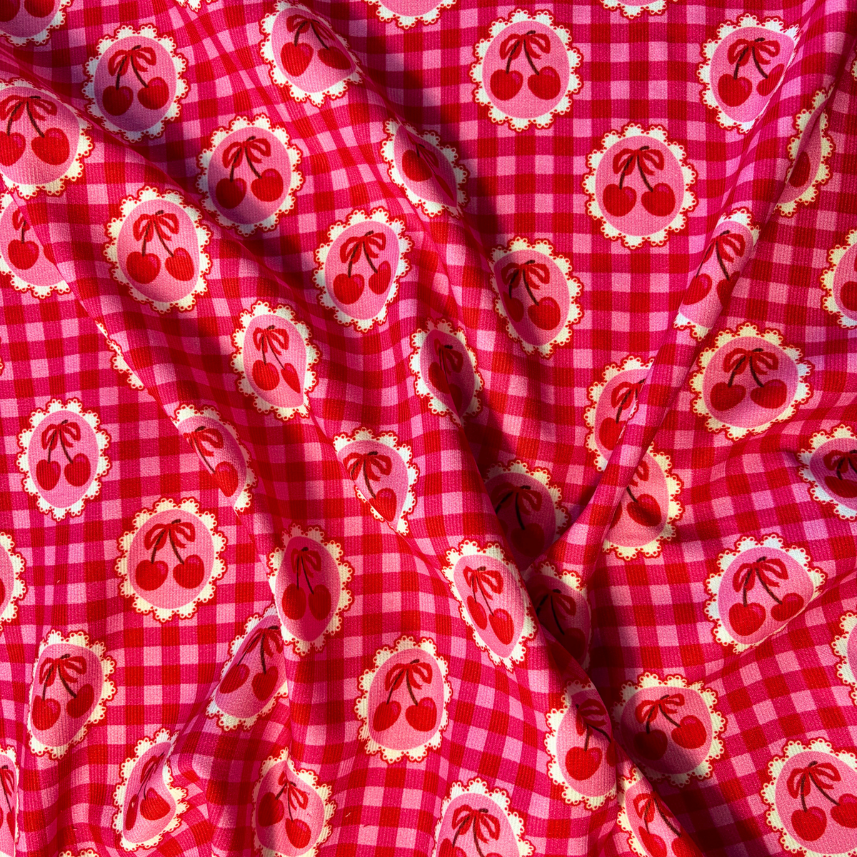 Red and pink gingham cherry needlecord fabric folded to show soft corduroy ridges and circular cherry motifs, 112cm wide cotton corduroy for handmade skirts bags and soft furnishings