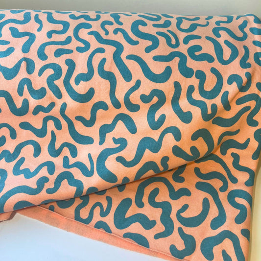 Flat lay of teal squiggle print sweatshirting fabric on peachy pink background, 285gsm brushed knit.