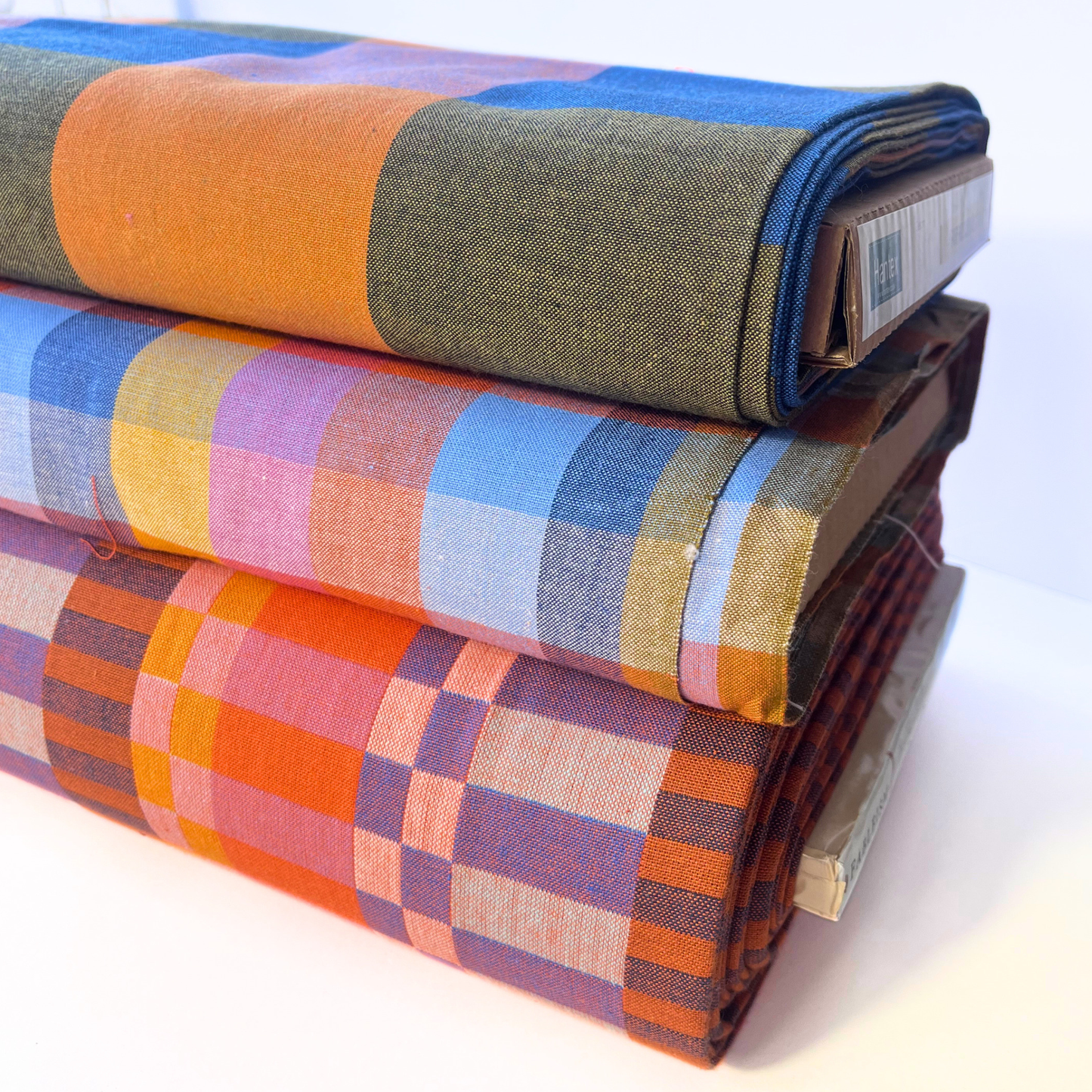 Three folded bolts of patterned fabric stacked on a white surface. The top fabric is olive green with a wide orange stripe and blue side panels.