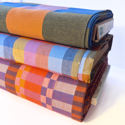 Three folded bolts of patterned fabric stacked on a white surface. The top fabric is olive green with a wide orange stripe and blue side panels.